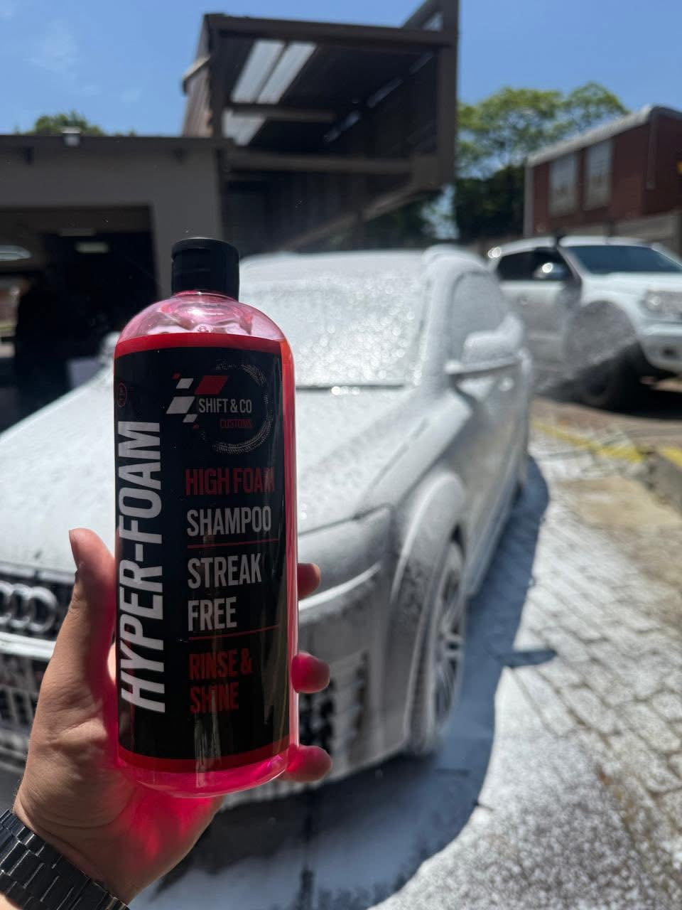 Person holding a bottle of 'Hyper-Foam' shampoo in front of a car being washed.